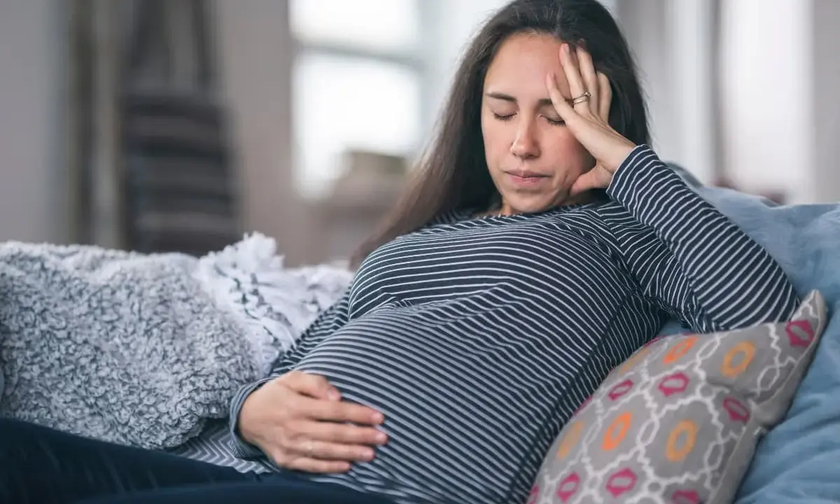 Gravid kvinna med ont i magen på natten, vilar på en soffa med handen på magen och pannan.