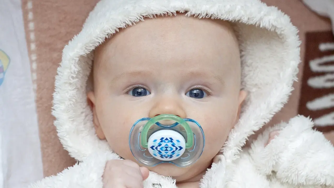 Söt bebis med blå ögon och en napp i munnen. Den fluffiga vita mössan ger ett mysigt intryck.