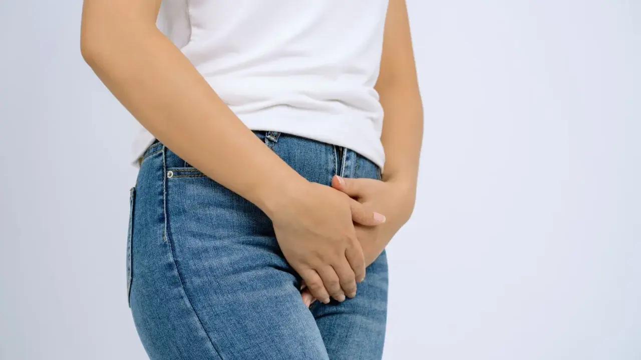 En person i jeans och vit t-shirt håller handen över underlivet, möjligen på grund av åderbråck i underlivet.