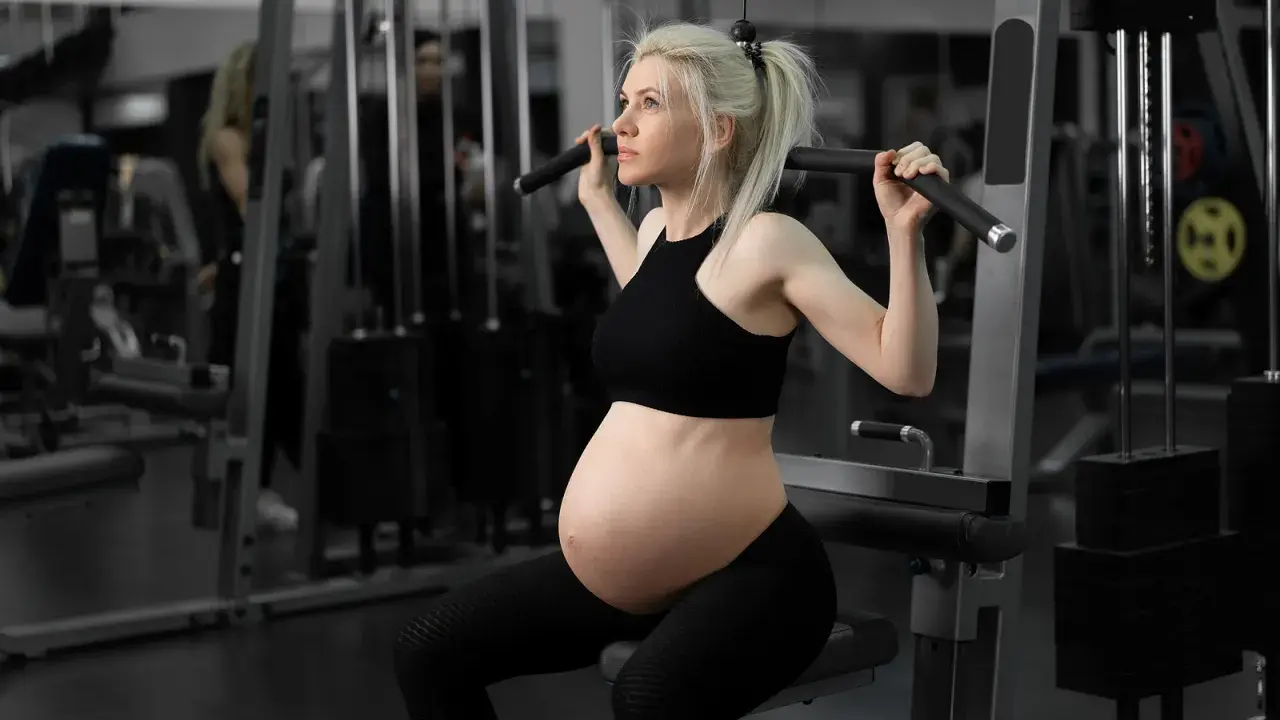 En **spinning gravid** tränar på gym. Hon sitter på en träningsmaskin och drar i ett handtag.