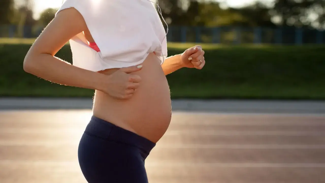 En gravid kvinna springer på en löparbana i solnedgången, med hörlurar i öronen.