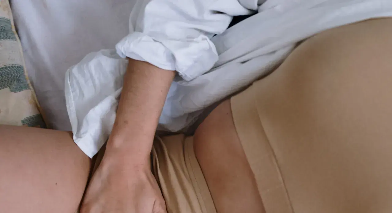 En person ligger i sängen, med en vit skjorta och hudfärgade underkläder. En hand vilar på låret, nära en slapp vagina.