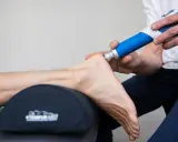 Behandling av plantar fasciit med stötvågsbehandling. En terapeut använder en blå enhet för att behandla en fot.