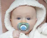 Söt bebis med blå ögon och en napp i munnen. Den fluffiga vita mössan ger ett mysigt intryck.