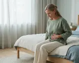 Kvinna med handen på magen sitter på sängkanten, kanske upplever hon biverkningar av en prolapsring.