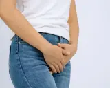 En person i jeans och vit t-shirt håller handen över underlivet, möjligen på grund av åderbråck i underlivet.