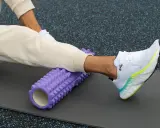 Person använder en lila foam roller rygg för att stretcha benet på en träningsmatta.