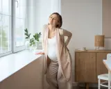 Gravid kvinna med ont i svanskotan lutar sig mot ett fönster. Hon håller handen på ryggen och grimaserar av smärta.