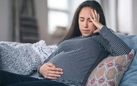 Gravid kvinna med ont i magen på natten, vilar på en soffa med handen på magen och pannan.