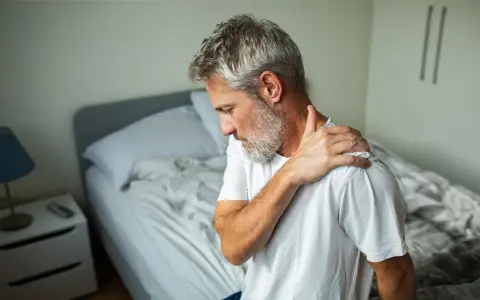 Man med grått hår sitter på sängkanten och håller sig om axeln, uppenbart med ont i nacken som strålar ut i armen.