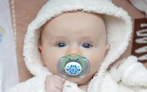 Söt bebis med blå ögon och en napp i munnen. Den fluffiga vita mössan ger ett mysigt intryck.