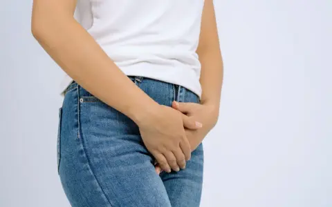 En person i jeans och vit t-shirt håller handen över underlivet, möjligen på grund av åderbråck i underlivet.