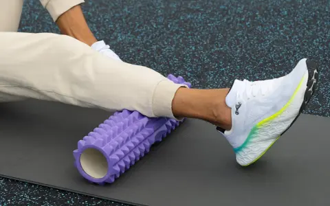 Person använder en lila foam roller rygg för att stretcha benet på en träningsmatta.
