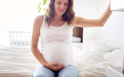 Gravid kvinna med smärta i magen, kanske upplever hon braxton hicks-sammandragningar. Hon sitter på en säng och håller sig om magen.