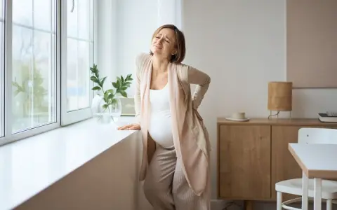 Gravid kvinna med ont i svanskotan lutar sig mot ett fönster. Hon håller handen på ryggen och grimaserar av smärta.