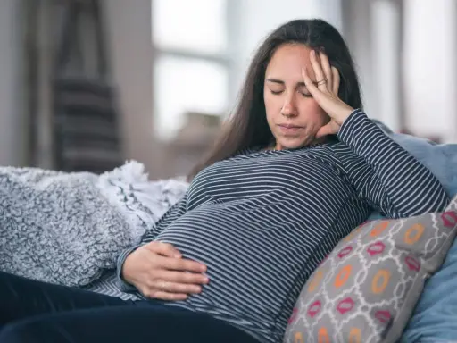 Gravid kvinna med ont i magen på natten, vilar på en soffa med handen på magen och pannan.