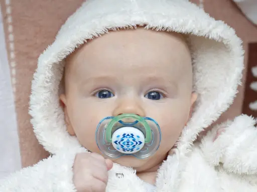 Söt bebis med blå ögon och en napp i munnen. Den fluffiga vita mössan ger ett mysigt intryck.