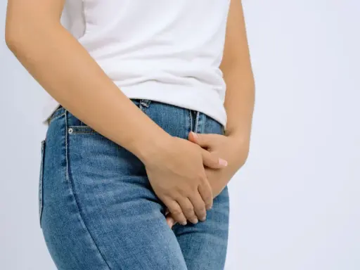 En person i jeans och vit t-shirt håller handen över underlivet, möjligen på grund av åderbråck i underlivet.