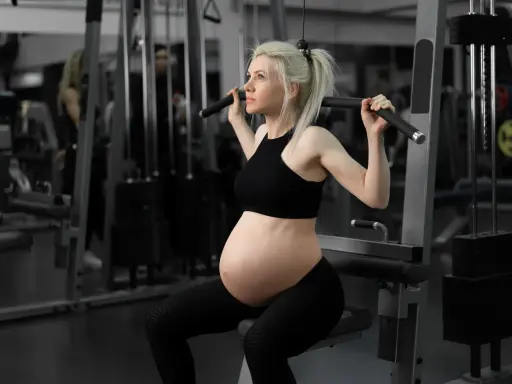 En **spinning gravid** tränar på gym. Hon sitter på en träningsmaskin och drar i ett handtag.