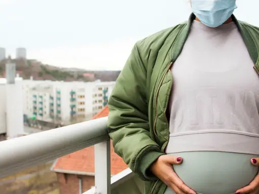 Gravid kvinna med munskydd på balkongen, omgiven av stadens silhuett. Känns det som att du är oftare sjuk gravid?