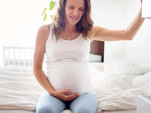 Gravid kvinna med smärta i magen, kanske upplever hon braxton hicks-sammandragningar. Hon sitter på en säng och håller sig om magen.