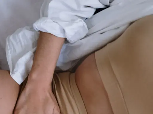 En person ligger i sängen, med en vit skjorta och hudfärgade underkläder. En hand vilar på låret, nära en slapp vagina.