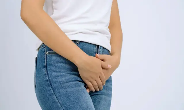 En person i jeans och vit t-shirt håller handen över underlivet, möjligen på grund av åderbråck i underlivet.