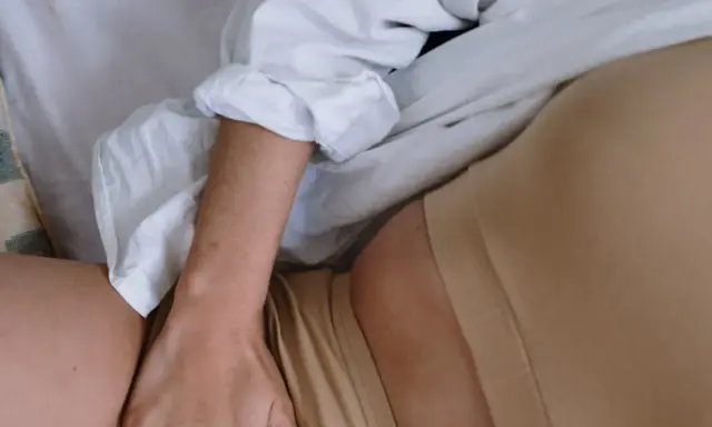 En person ligger i sängen, med en vit skjorta och hudfärgade underkläder. En hand vilar på låret, nära en slapp vagina.