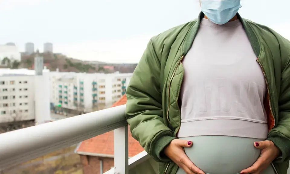 Gravid kvinna med munskydd på balkongen, omgiven av stadens silhuett. Känns det som att du är oftare sjuk gravid?
