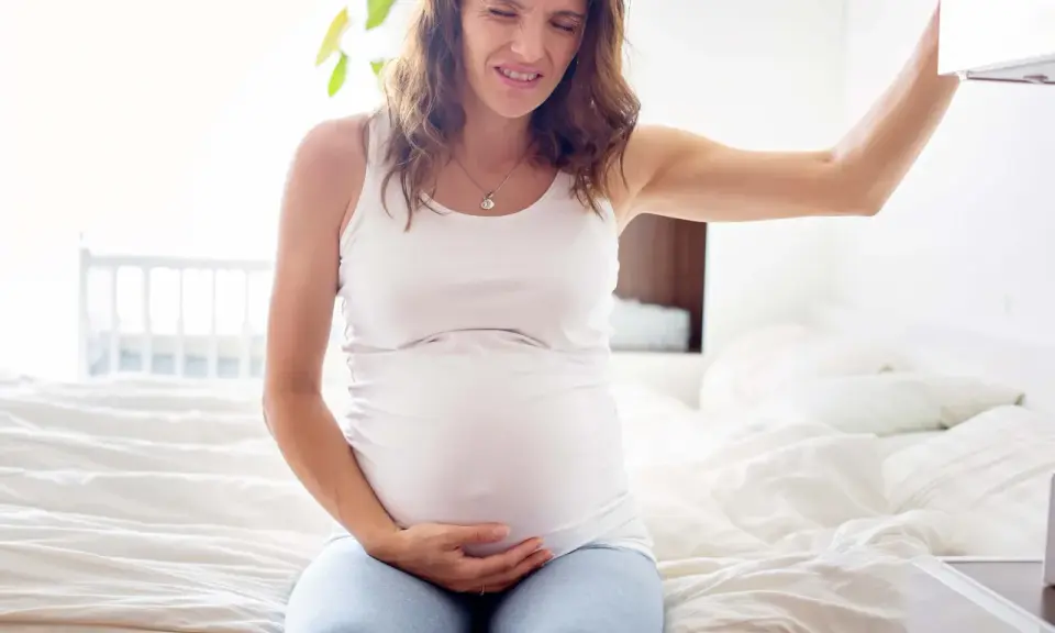 Gravid kvinna med smärta i magen, kanske upplever hon braxton hicks-sammandragningar. Hon sitter på en säng och håller sig om magen.