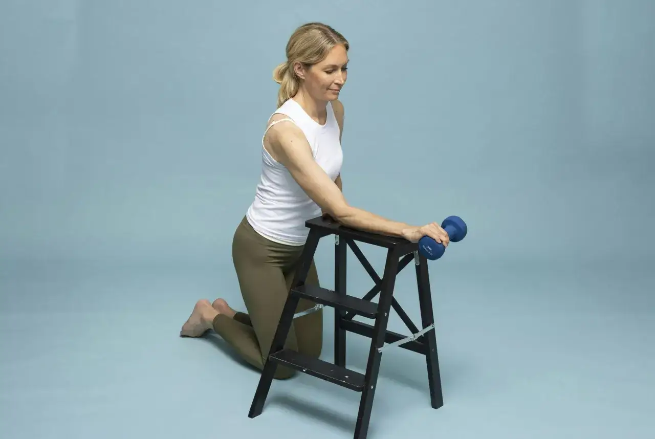 Kvinna med tennisarmb&aring;ge utf&ouml;r rehabiliteringstr&auml;ning med hantel p&aring; en pall.