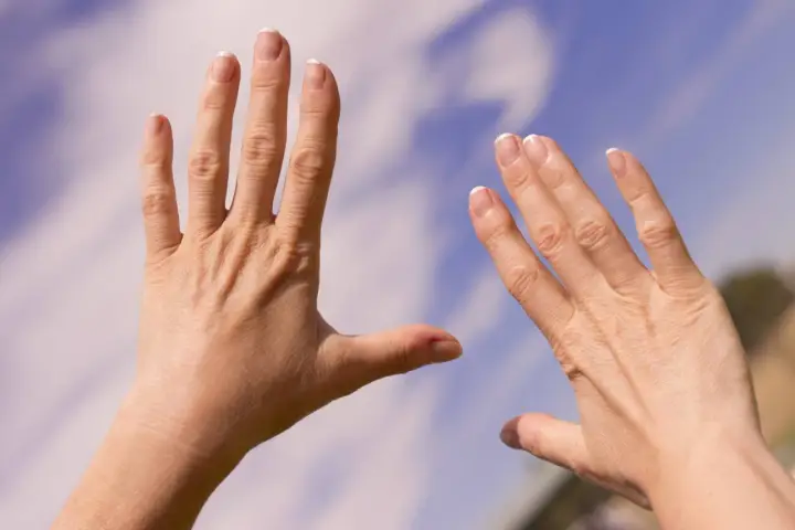 Två händer med sneda fingrar, kanske ett tecken på reumatism, sträcks mot en blå himmel med moln.